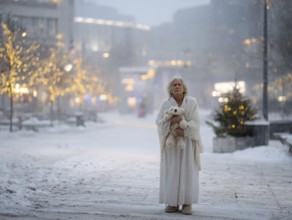 An elderly woman in a nightgown stands with her teddy bear in a cold, wintry environment, looking