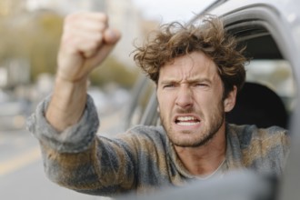 A man in a car shows a clenched fist and looks angrily out of the window on a street, symbolic
