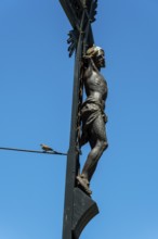A detailed sculpture depicting Jesus on a cross rises against a bright blue sky. A small bird can