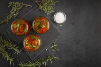 Three glasses filled with pepperoni and herbs on a dark surface next to salt
