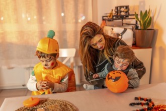 Mother and children wearing halloween costumes are sitting at a table at home, playing with pumpkin