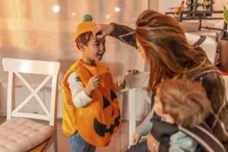 Mother applying halloween makeup on her son wearing a pumpkin costume, while holding her baby