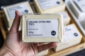 Hand holding firm tofu block in front of supermarket refrigerator. Concept of plant-based protein