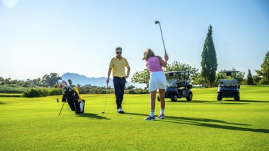 Golf instructor coaching female golfer on driving range, with golf carts in background on sunny day