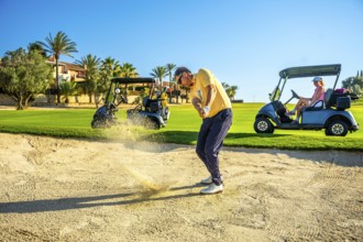 Golfer hitting ball out of sand trap with golf carts and other golfers in background on sunny day