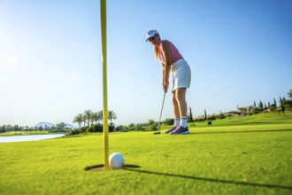 Woman focusing on a putt on a bright, sunny day at a lush golf course, with a ball near the hole