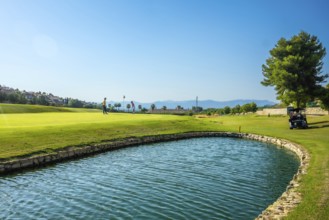 Two golfers are playing on a sunny golf course with a water hazard and golf cart in the background