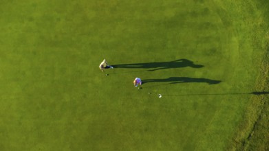 Two golfers are playing on a perfectly manicured green golf course on a sunny day, casting long