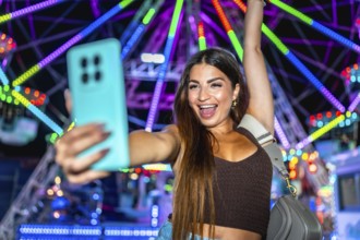 Young woman taking a selfie with smartphone in front of a colorful ferris wheel at an amusement