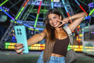 Happy young woman taking a selfie and making peace sign at amusement park with colorful ferris