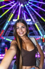 Happy young woman taking a selfie with a colorful ferris wheel in the background at an amusement