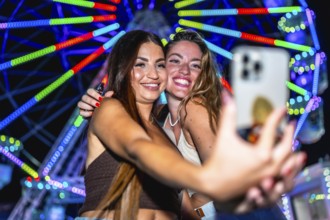 Two friends are taking a selfie in front of a brightly lit ferris wheel at an amusement park at