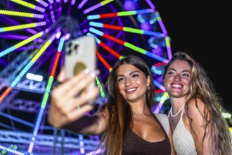 Two happy women taking a selfie with smartphone in front of illuminated ferris wheel at an