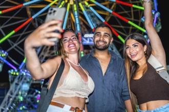 Three happy friends taking a selfie with smartphone in front of a colorful ferris wheel at an