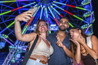 Three friends taking a selfie with a smartphone in front of a brightly lit ferris wheel at an