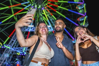 Three friends taking a selfie with smartphone in front of a colorful ferris wheel, enjoying