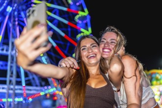 Two cheerful young women taking a selfie in front of a brightly lit ferris wheel at an amusement