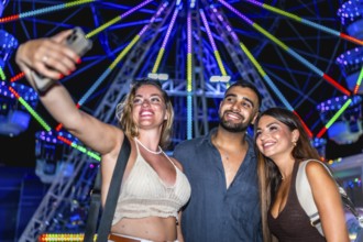 Three happy friends taking a selfie with smartphone in front of a colorful ferris wheel at an
