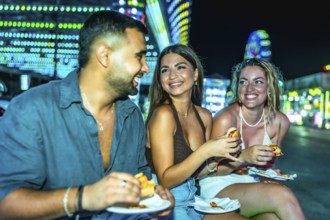 Happy friends sharing delicious hot dogs while enjoying the vibrant atmosphere of an amusement park