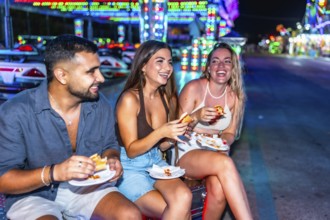 Three friends are sitting and enjoying hot dogs together at an amusement park, illuminated by