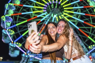 Two young women taking a selfie with a smartphone in front of a brightly lit ferris wheel at an