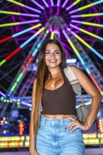 Happy young woman posing at night with illuminated ferris wheel in background, enjoying funfair