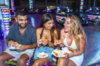 Three friends are sitting and laughing while eating hot dogs at an amusement park's bumper cars
