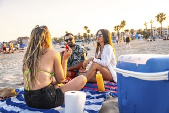Three friends are sitting on a beach towel, enjoying a picnic with watermelon and drinks at sunset