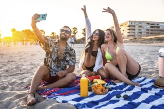 Three friends are sitting on a beach towel, taking a selfie while enjoying snacks and drinks during