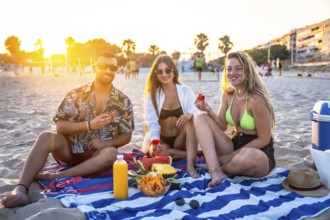 Three friends enjoying a picnic on the beach at sunset, eating watermelon and other fresh fruit