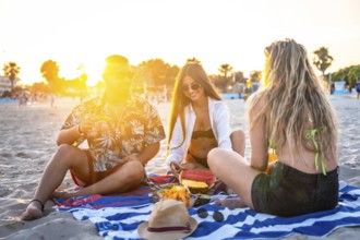 Group of friends enjoying watermelon and pineapple during a sunset picnic on a beach towel,