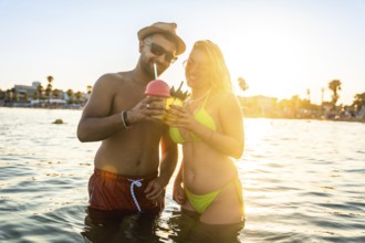Happy couple enjoying refreshing cocktails while standing in the ocean during a beautiful sunset