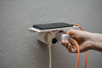 A person is holding a charger connector and plugging it into a wall-mounted power outlet. A