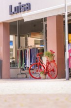 Red bicycle with flower baskets in front of a clothing shop in a busy shopping street, small town