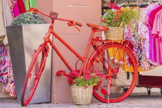 A decorated red bicycle stands in front of a clothing shop with colourful clothes, small town of