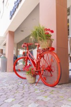 A red bicycle with flower baskets stands decoratively on a pavement in front of shops, small town