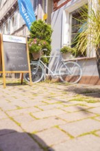Bicycle with flower baskets and chalkboard in front of a café in the sunshine, small town of Perle