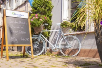 A pale bicycle with flowers in baskets next to a chalkboard in front of a café, small town of Perle