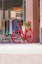 Bright red bicycle with flower arrangement in front of a fashion shop in a sunny street, small town