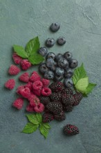 A mix of ripe berries, with leaves scattered on the table, raspberries, blackberries, blueberries,