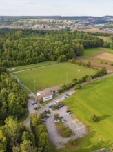 Aerial view of a sports field in the middle of wooded and rural surroundings with parked cars and a