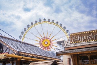 Large Ferris wheel behind wooden buildings, in a bright and cloudy sky, Olympiapark, Munich,