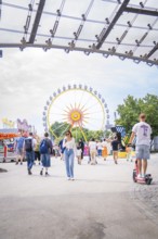People in amusement park with e-scooter and big Ferris wheel in the background, Olympiapark,