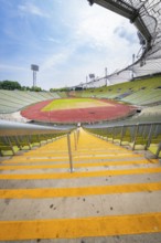 View down the yellow steps to an empty stadium with wide stands and running track, Olympiapark,