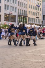 Dance group performs outdoors against an urban backdrop, surrounded by spectators, Munich, Germany