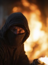 Vermummte Person mit Kapuze und Maske hält Flasche vor einem großen Feuer, Brandstiftung,