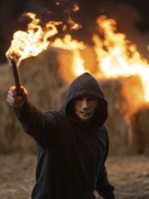 Maskierte Person hebt Fackel vor brennendem Strohlager in der Nacht, Brandstiftung, Brandleger,