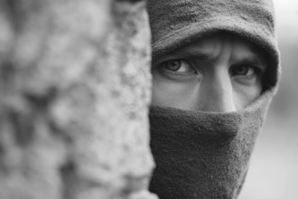 Eine vermummte Person schaut mit intensivem Blick hinter einer Mauer und beobachtet eine Situation,