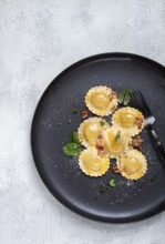 Fresh ravioli, with butter, spices and herbs, fried bacon, on a black plate, top view, dark