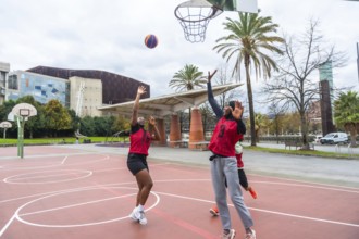 Two diverse women in red jerseys actively playing basketball on an outdoor court. Reaching for the